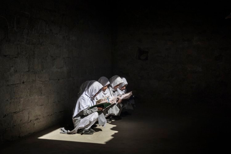 Students in Pakistan sitting in sunlight from a window to read books in a dark room.